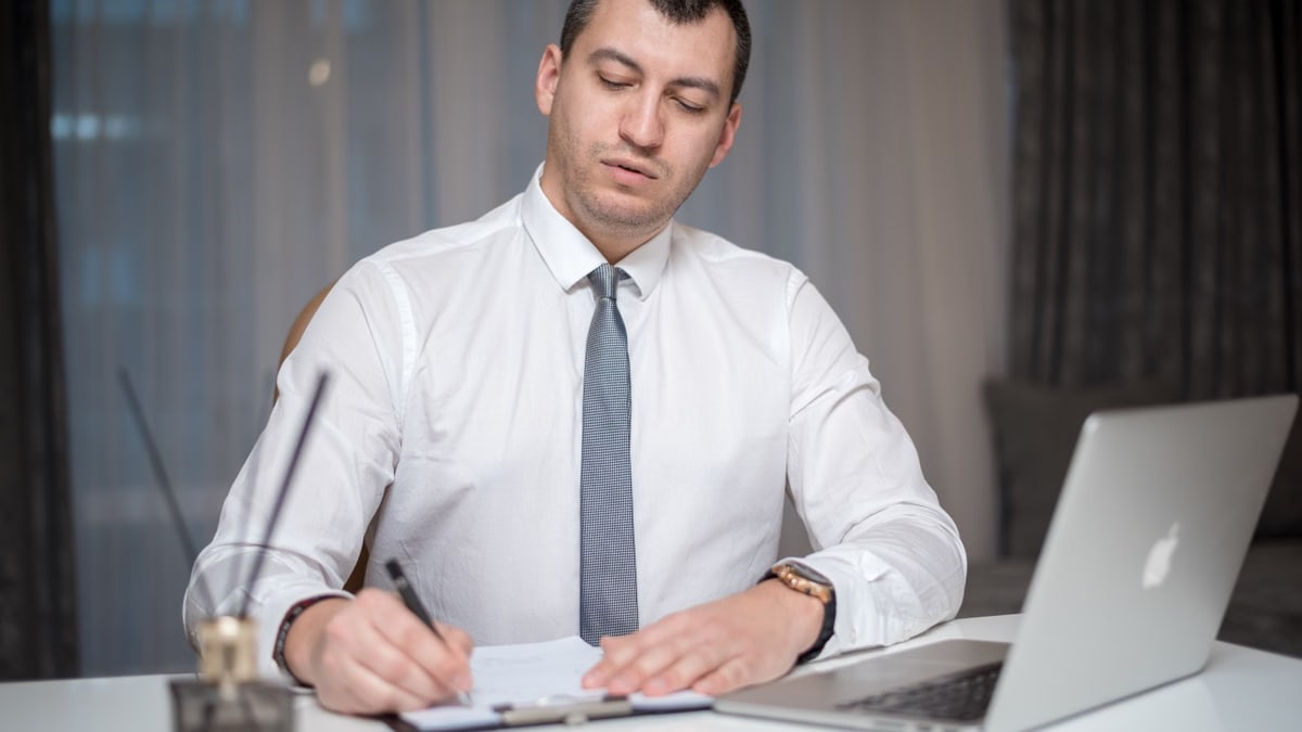 Focused businessman in office writing notes on paper with laptop at a white desk.
