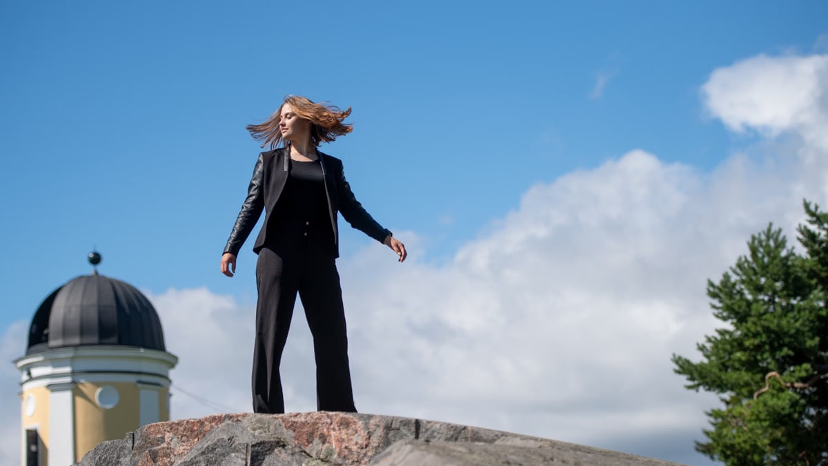 Young woman posing confidently on a rock in a Helsinki park, with a clear blue sky and European architecture in the background.